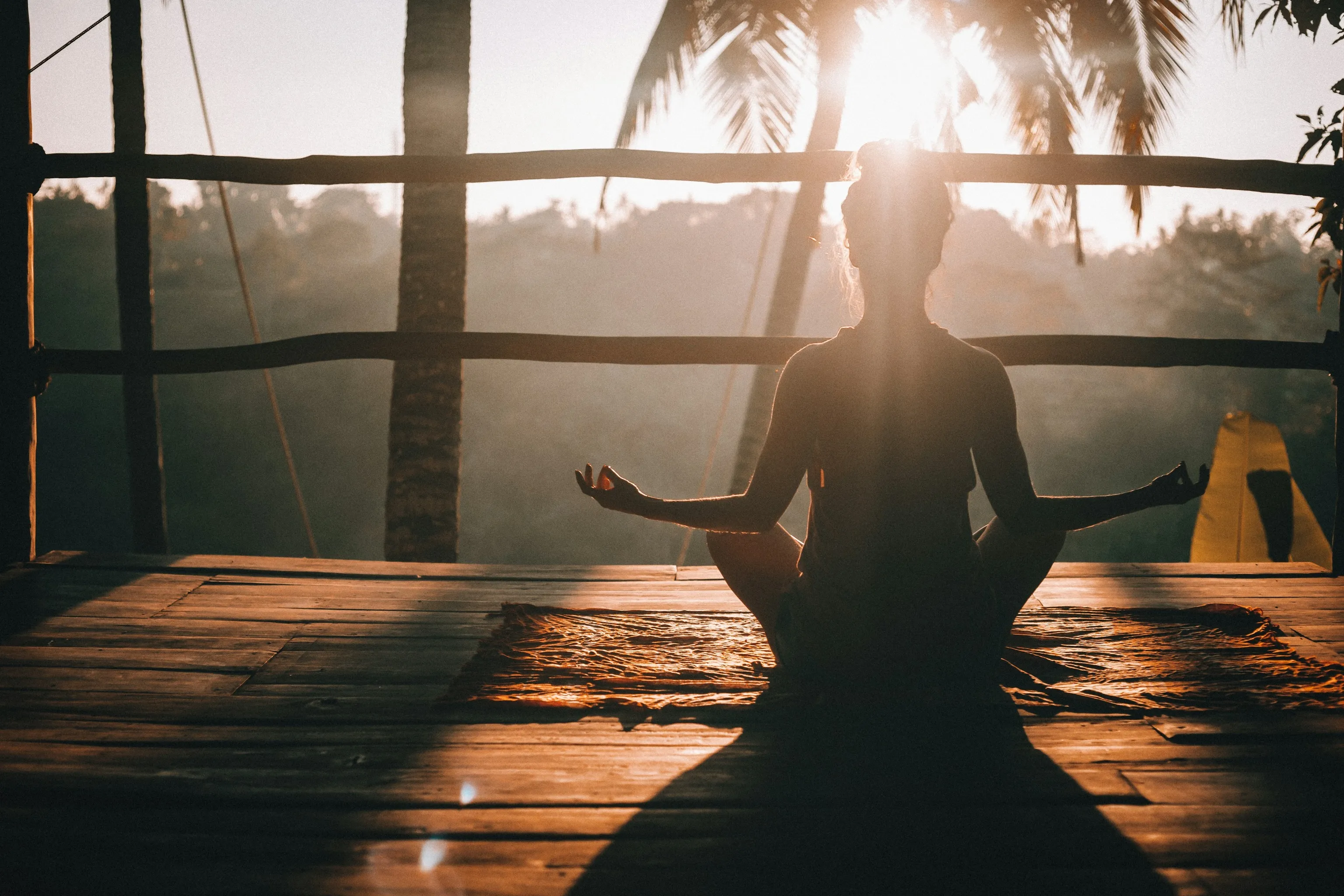 Woman meditating cross-legged on a timber deck at sunrise, framed by palms